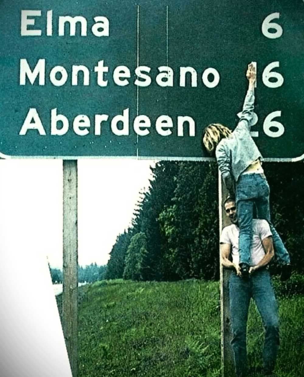 A person stands on anothers shoulders, reaching up to touch a large green road sign listing Elma, Montesano, and Aberdeen, each 6 miles away, beside a grassy roadside and trees.