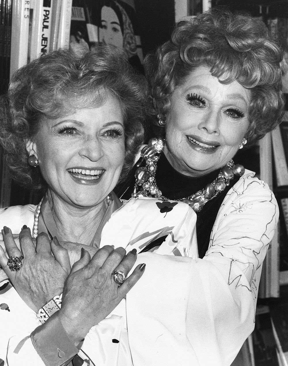 Betty White and Lucille Ball with curly hair pose together. One woman stands behind the other with her arms affectionately around her. Both wear patterned outfits and jewelry. Books and posters are visible in the background.