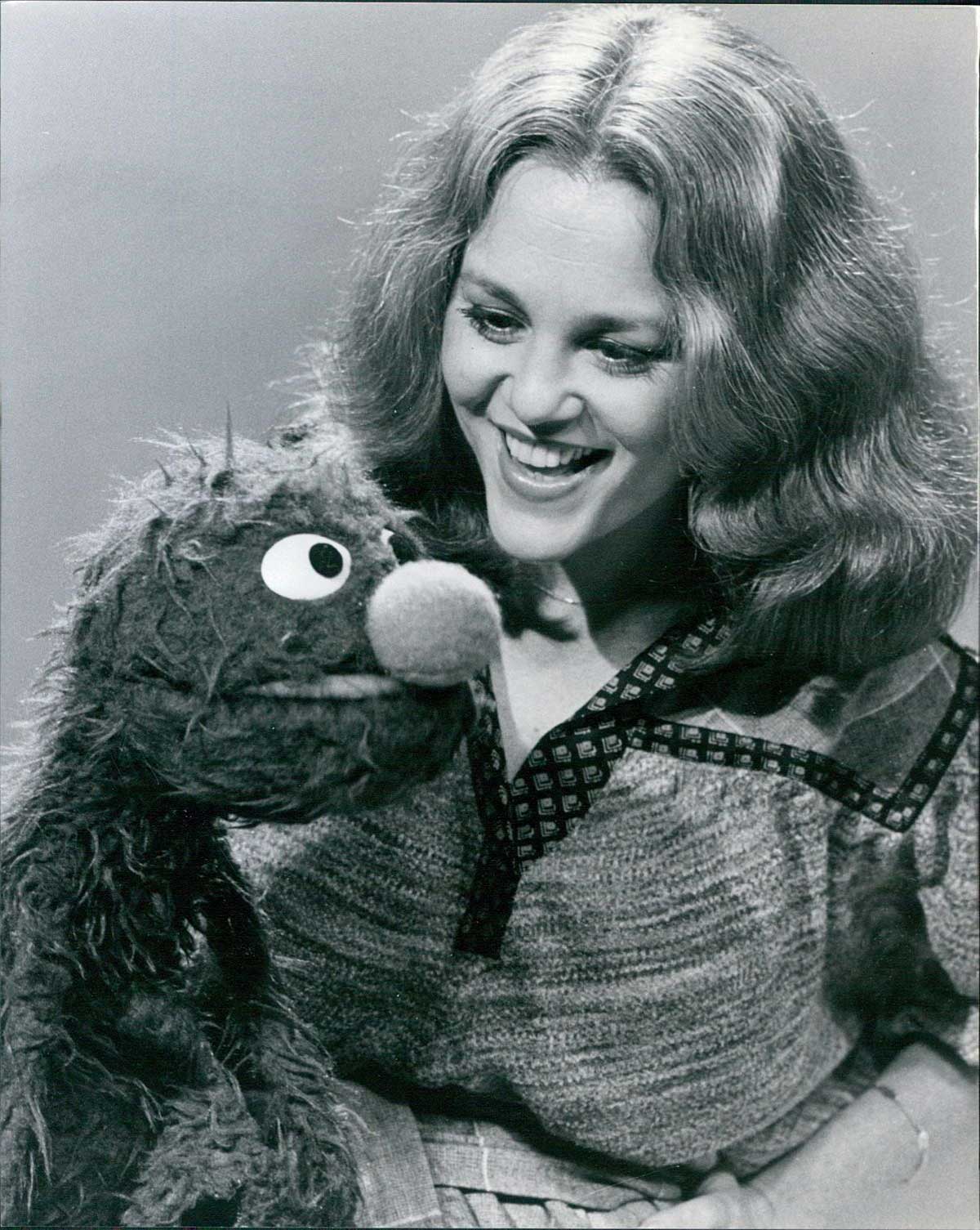 A woman with wavy hair smiles at a furry puppet with a round nose and big eyes. The image is black and white, and they appear to be engaged in a joyful interaction.