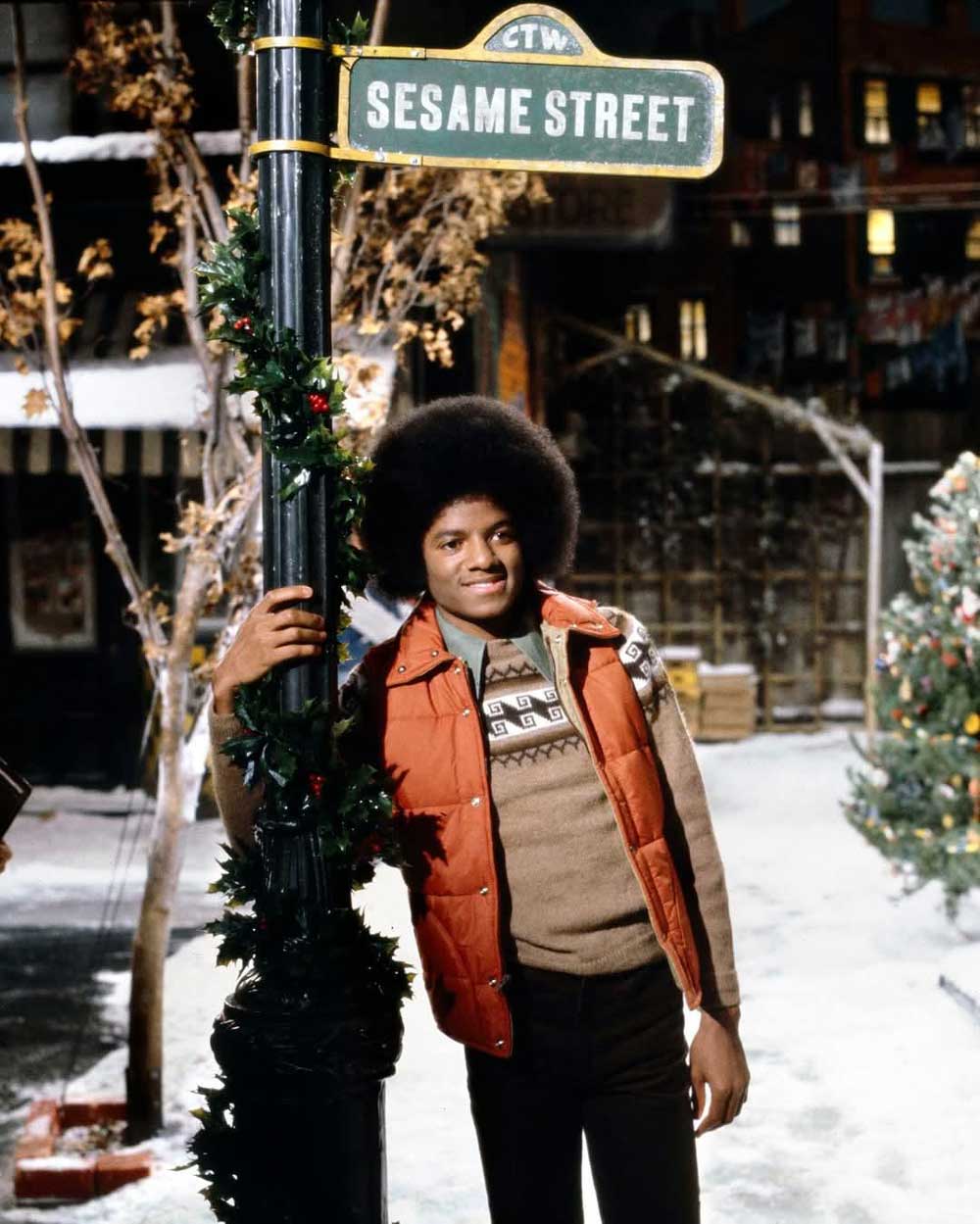 A young man in a red vest and brown sweater smiles, leaning against a lamppost decorated with garland under a Sesame Street sign on a snowy set with trees and holiday decorations in the background.