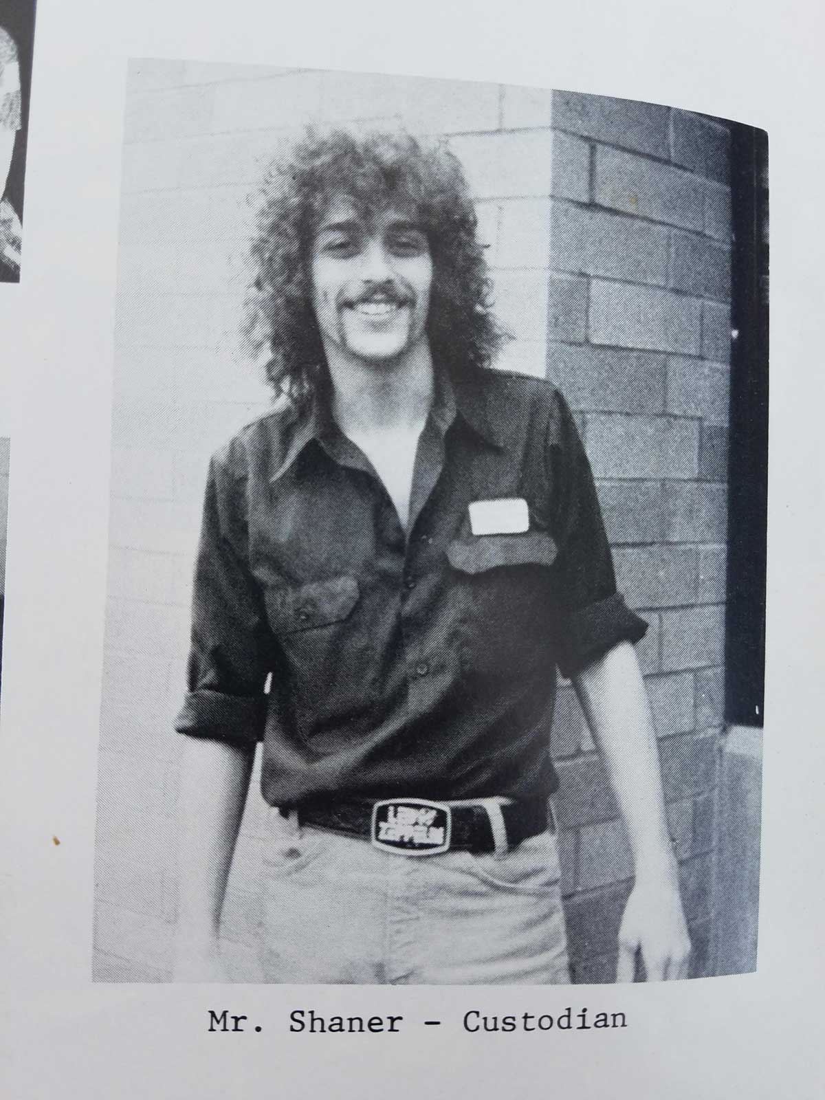 Black and white photo of a man with curly hair and a mustache, wearing a dark button-up shirt and a belt, smiling while standing in front of a brick wall. Text below reads: Mr. Shaner – Custodian.
