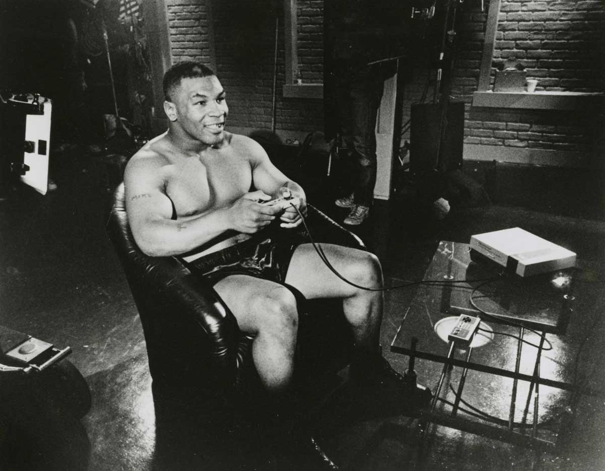 Mike Tyson in boxing shorts sits on a leather chair, smiling and playing a Nintendo Entertainment System console in a room with brick walls and studio equipment.