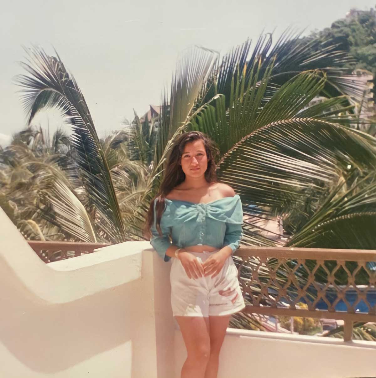 A woman with long hair stands on a balcony in front of palm trees, wearing a light blue off-shoulder top and white shorts. Its a sunny day with greenery and a hillside in the background.