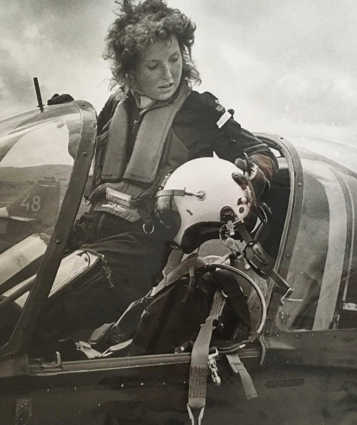 A woman in a flight suit stands beside an open cockpit of a jet, holding a pilot helmet and looking inside the aircraft. The image is in black and white, and the cockpit details and harness are visible.