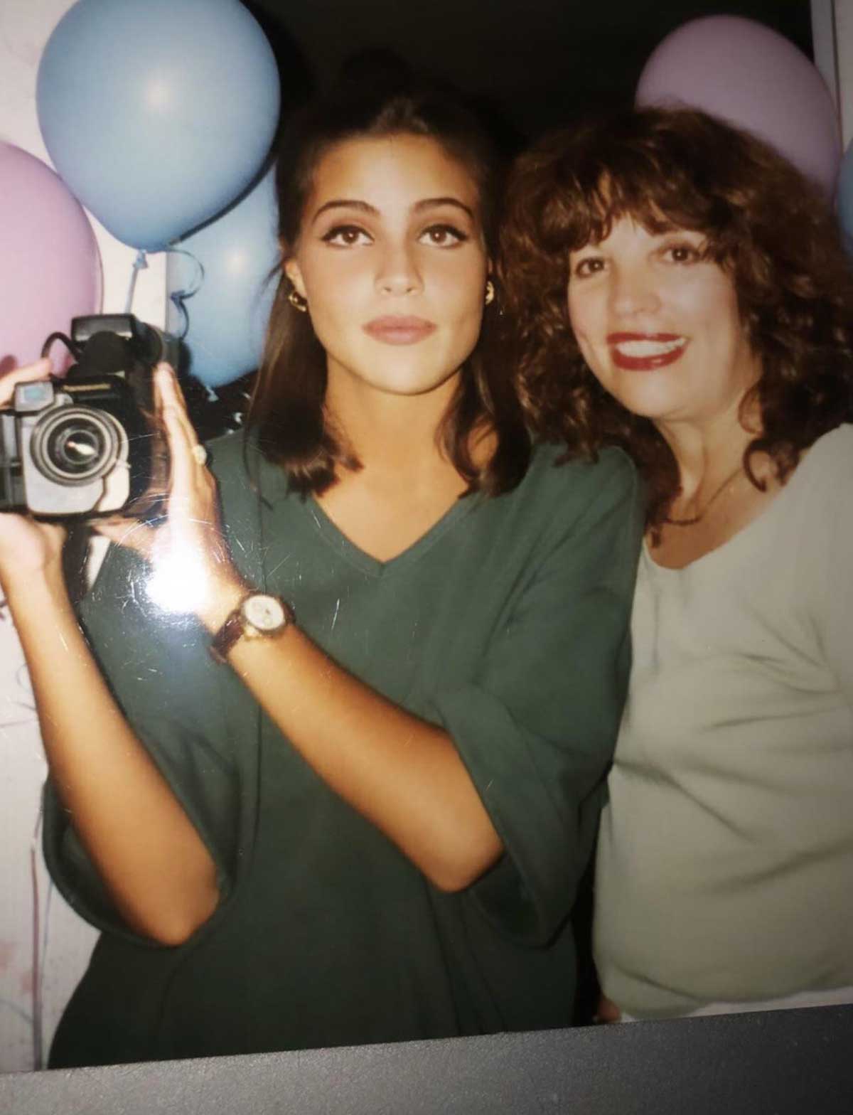 Two women pose closely together, one holding a camera, surrounded by pastel blue and pink balloons. Both are smiling softly, dressed casually, and appear to be at a celebratory event.