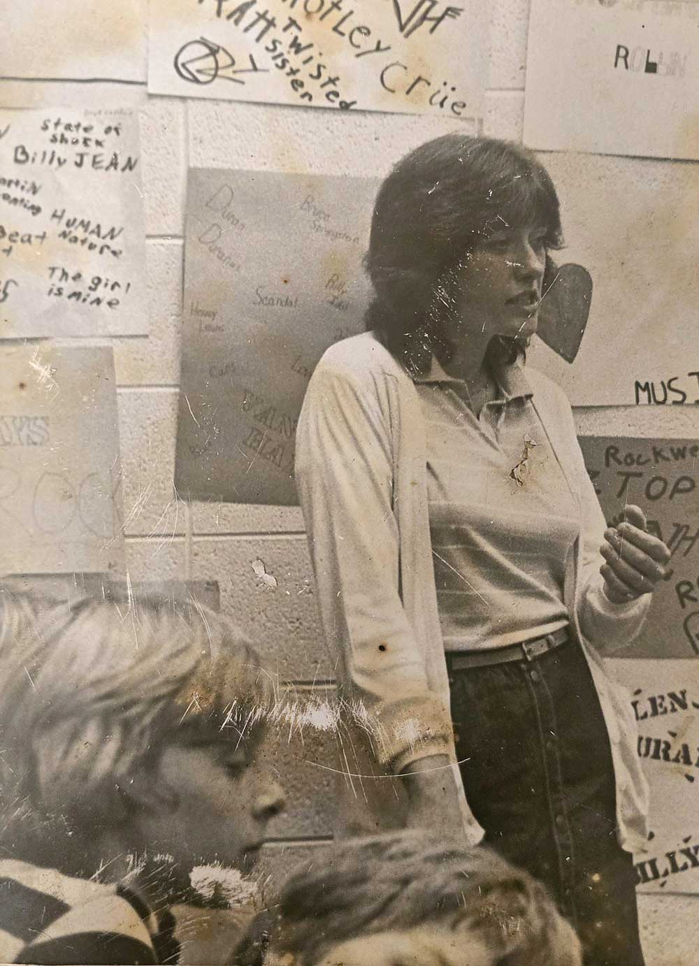 A woman stands in front of a classroom wall covered with handwritten posters. She wears a light cardigan over a collared shirt. Two children sit in the foreground, one partially visible. The scene appears vintage.