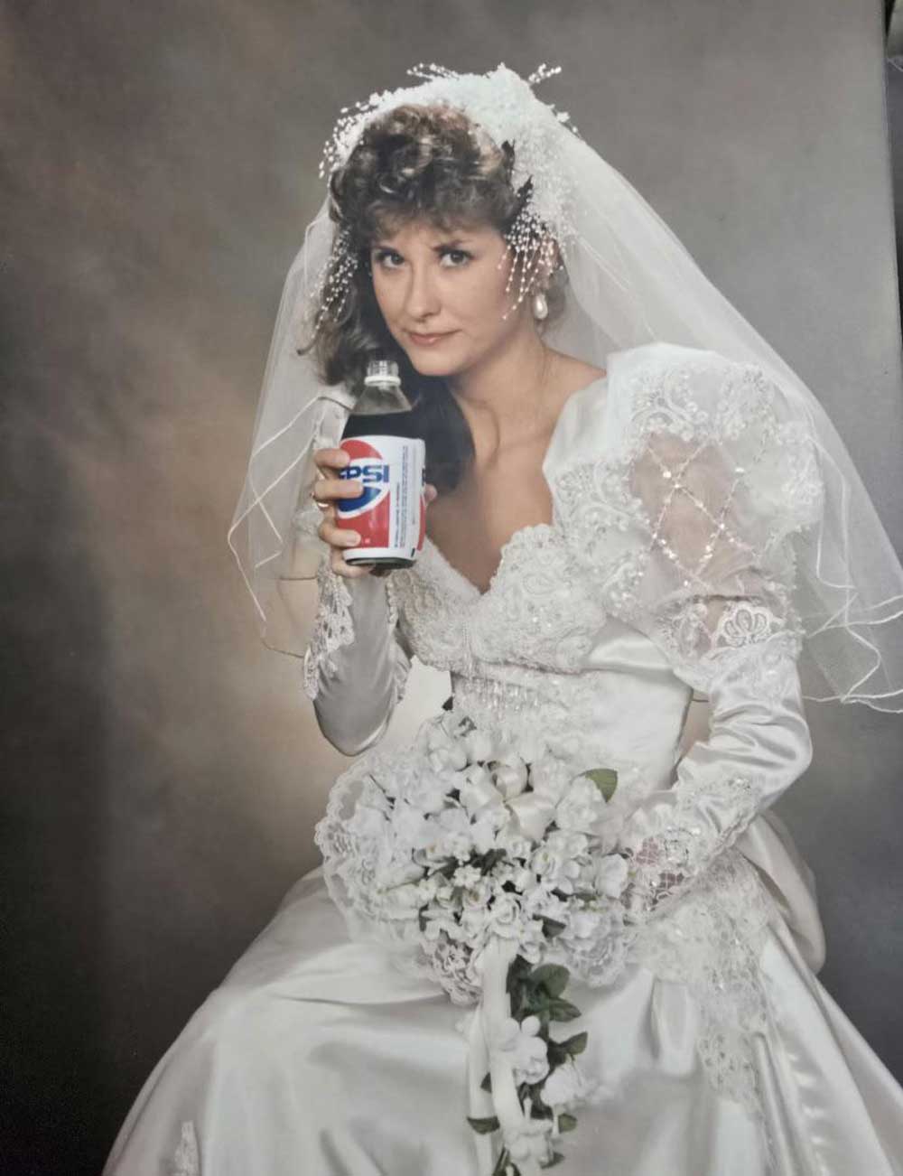 A bride in a white wedding dress and veil holds a bouquet of white flowers and a can of Pepsi, looking at the camera with a slight smile. The background is a classic studio portrait setting.