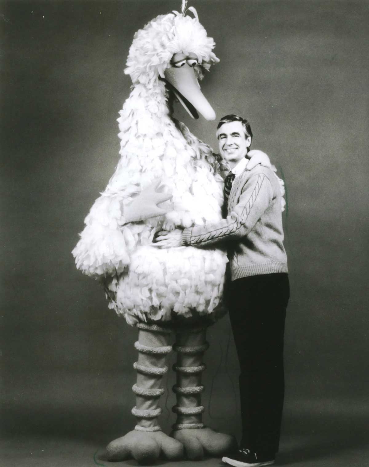 A man in a sweater smiles as he hugs Big Bird, a large, feathered character from Sesame Street, in a black and white studio photo.