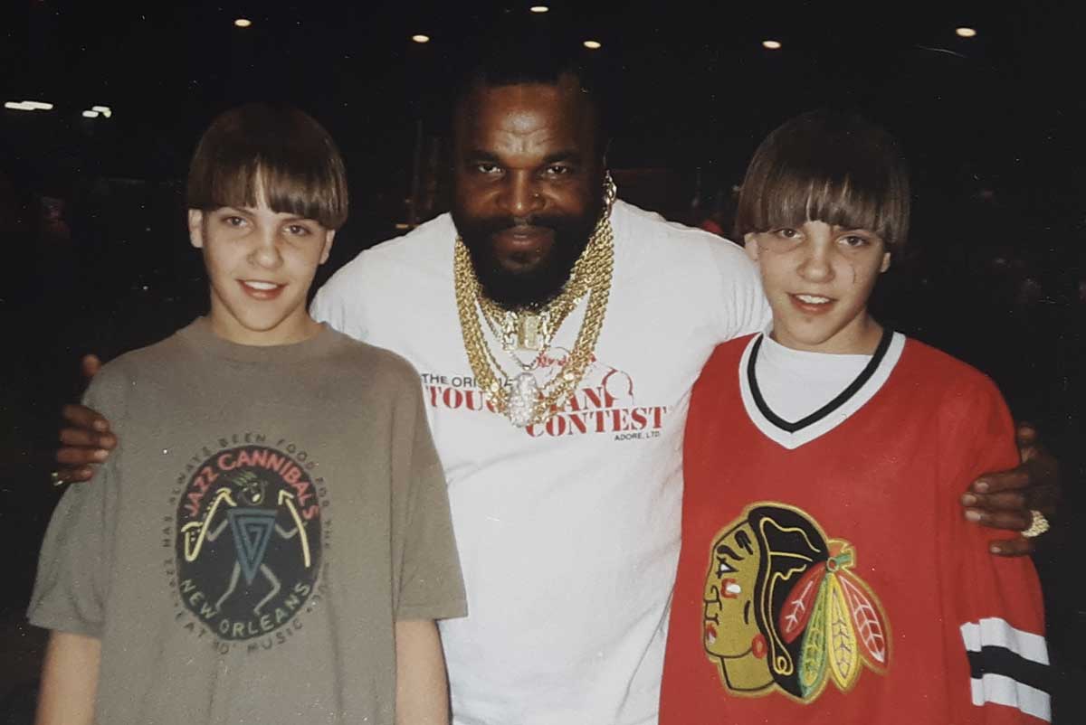 A man with a beard and gold chains stands smiling with his arms around two boys. One boy wears a graphic T-shirt and the other wears a red Chicago Blackhawks jersey. They are indoors with dim lighting in the background.