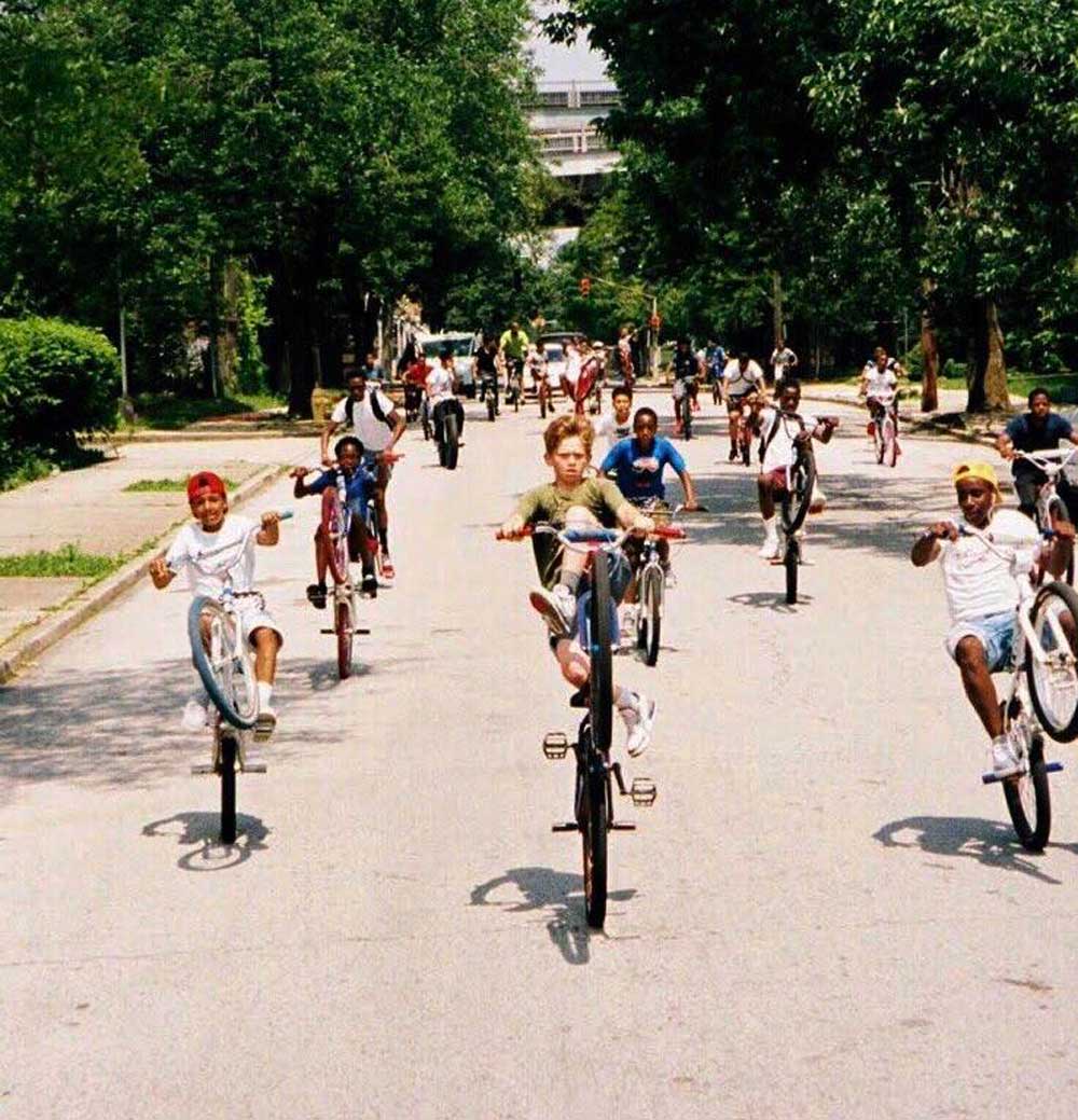 A group of kids ride bicycles down a sunny street, many performing wheelies. The scene is lively, with trees lining the road and more children biking in the background.