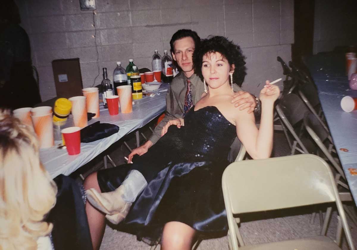 A woman in a black dress and boots sits on a mans lap at a table with drinks and cups. She holds a cigarette and looks at the camera, while the man, dressed in a shirt and tie, sits behind her. Party scene.