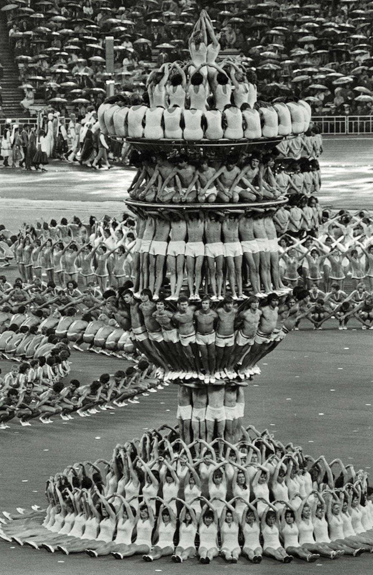 A large group of performers in matching outfits create an intricate, layered human formation resembling a vase or chalice during a stadium event, with more performers in synchronized poses in the background.