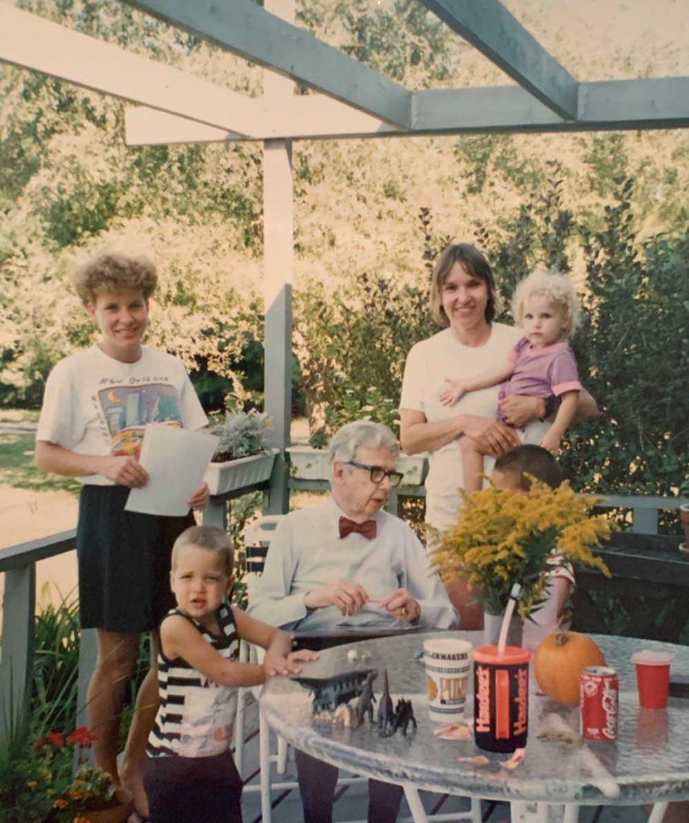 Five people, including two children and an elderly man with glasses and a bow tie, gather around a patio table outdoors. The table has drinks, flowers, toy dinosaurs, and a pumpkin. Green trees fill the background.