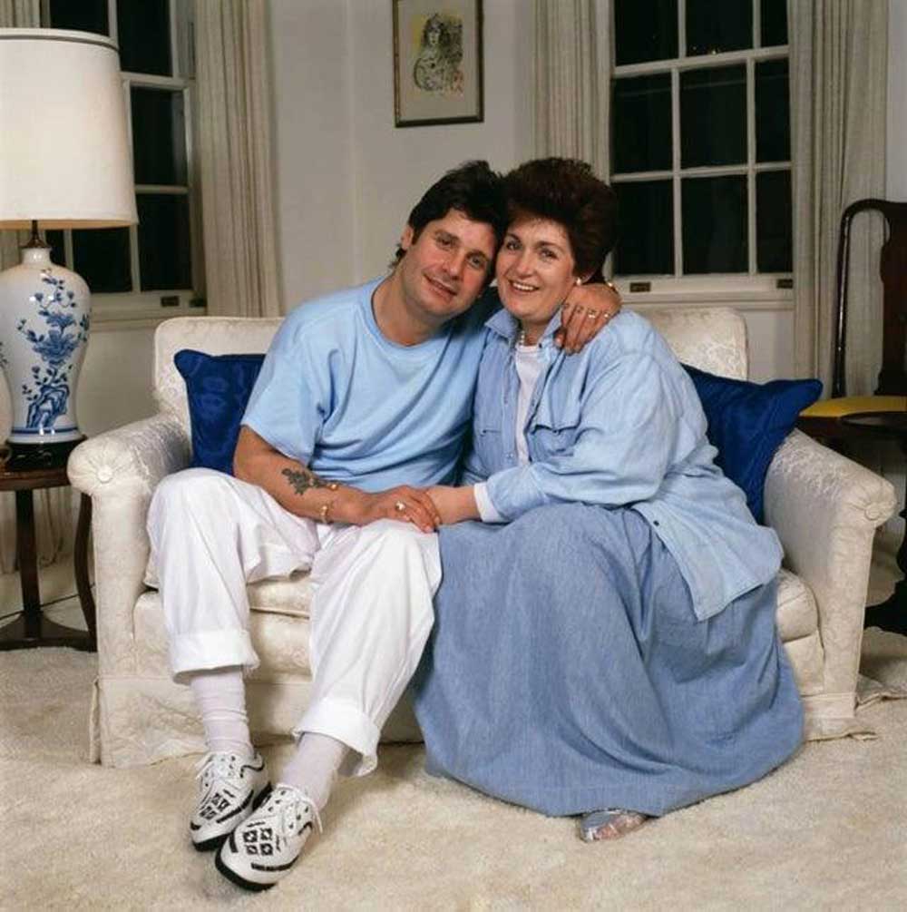 Ozzy and Sharon Osbourne sit closely together on a white couch, smiling at the camera. The man wears a light blue shirt and white pants; the woman wears a long blue dress. The room has white walls and a lamp on a side table.