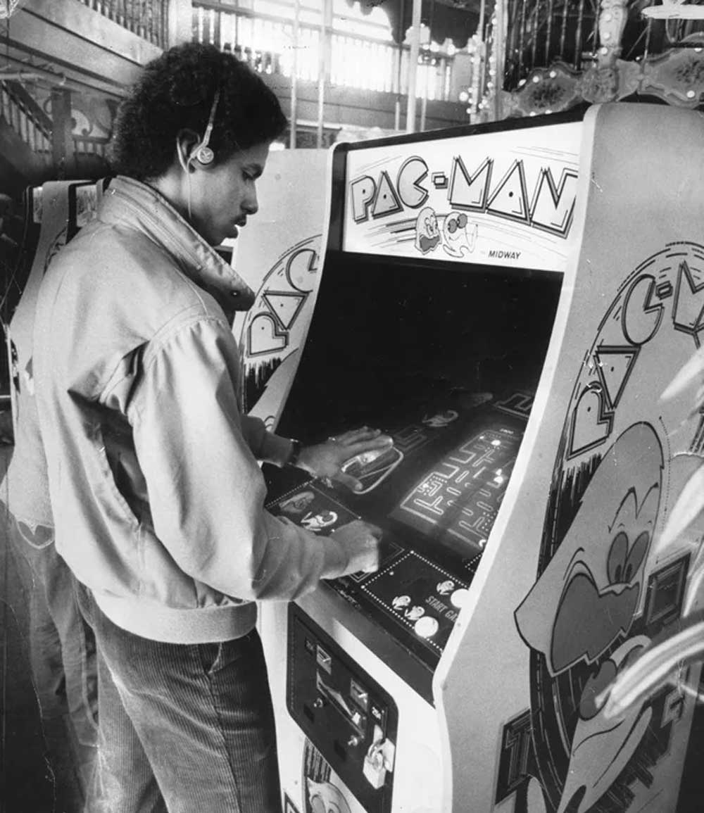 A person wearing headphones plays a Pac-Man arcade game in an indoor setting, possibly an arcade, with another arcade machine and decorative lights visible in the background.