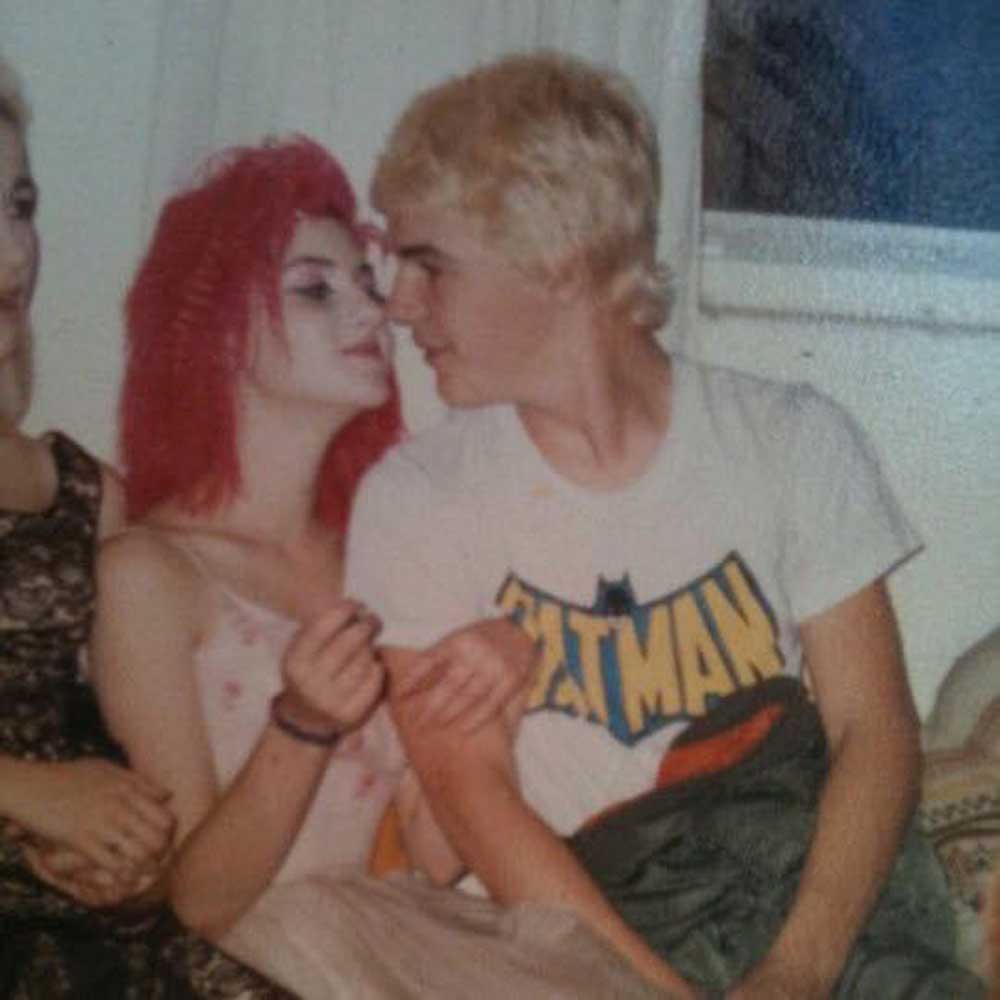 A person with bright red hair and dramatic makeup leans in closely to another person with short blond hair wearing a white Batman T-shirt; they appear to be gazing at each other affectionately.