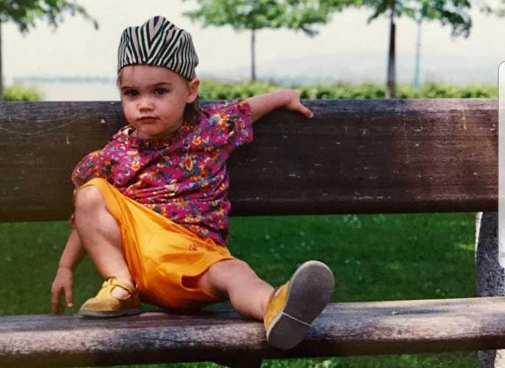 A young child wearing a striped hat, colorful floral top, and yellow shorts sits casually on a wooden bench outdoors, with one arm draped over the backrest and legs stretched out. Trees and green grass are in the background.