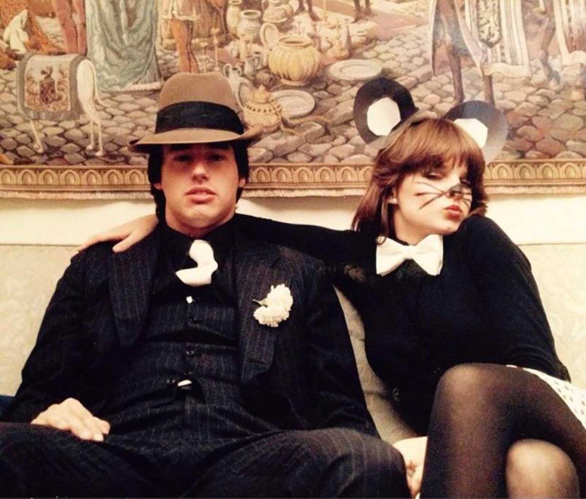 A man in a pinstripe suit and fedora sits next to a woman dressed as a mouse, with drawn-on whiskers, black ears, and a bow tie. They are sitting on a couch in front of a detailed, medieval-style tapestry.