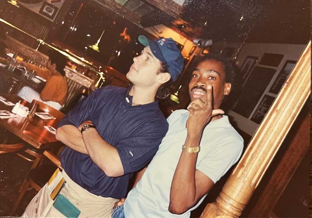 Two men posing in a casual restaurant. One, wearing a blue shirt and backward cap, stands with arms crossed. The other man, in a light shirt, points at the camera and smiles. Wooden decor and tables are visible in the background.