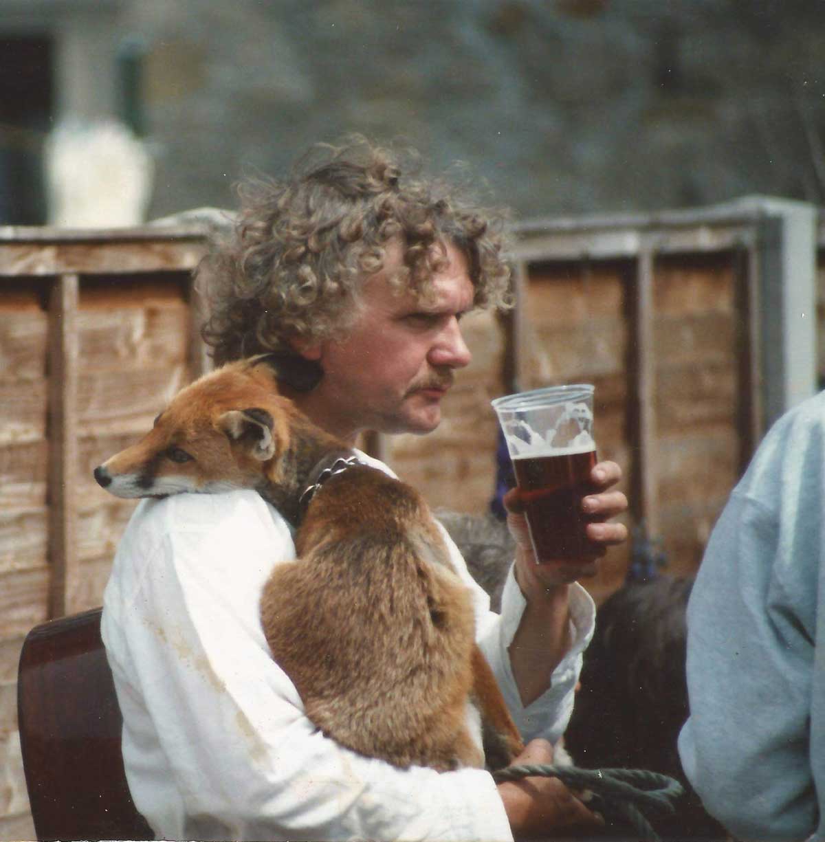A man with curly hair holds a pint of beer in one hand and a fox in the other arm while sitting outdoors near a wooden fence.