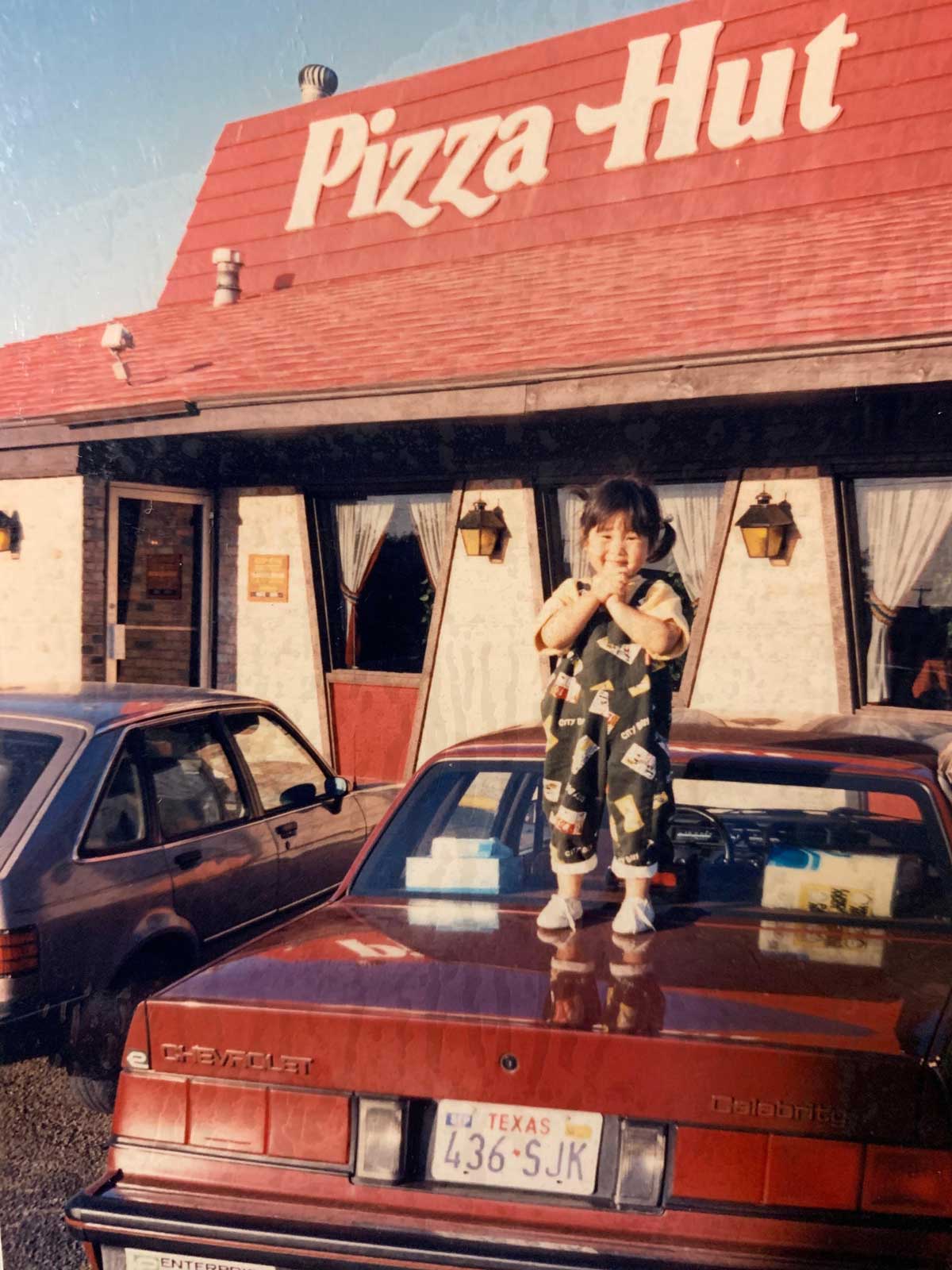 A young child stands on the hood of a red Chevrolet parked outside a Pizza Hut, smiling and giving a thumbs up. The restaurants name is visible on the red roof. Another car is parked nearby.