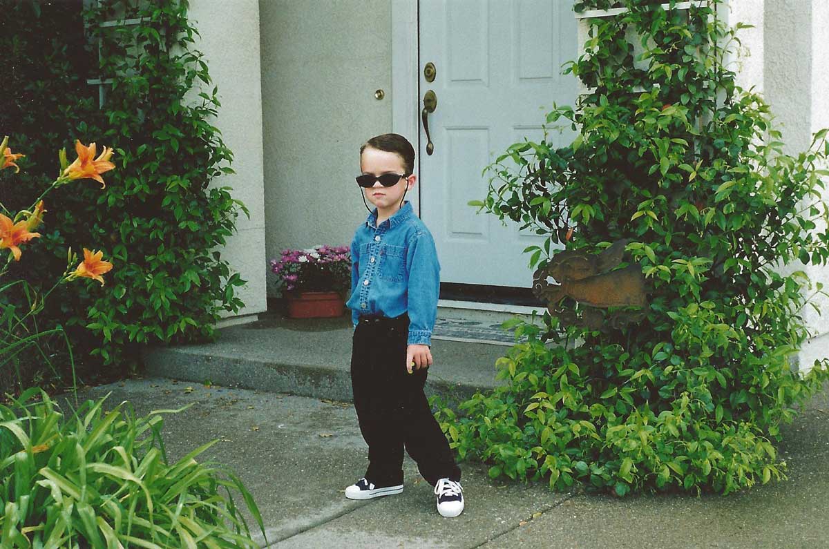 A young boy wearing sunglasses, a denim shirt, black pants, and sneakers stands confidently on a sidewalk in front of a house with greenery and orange flowers around the entrance.