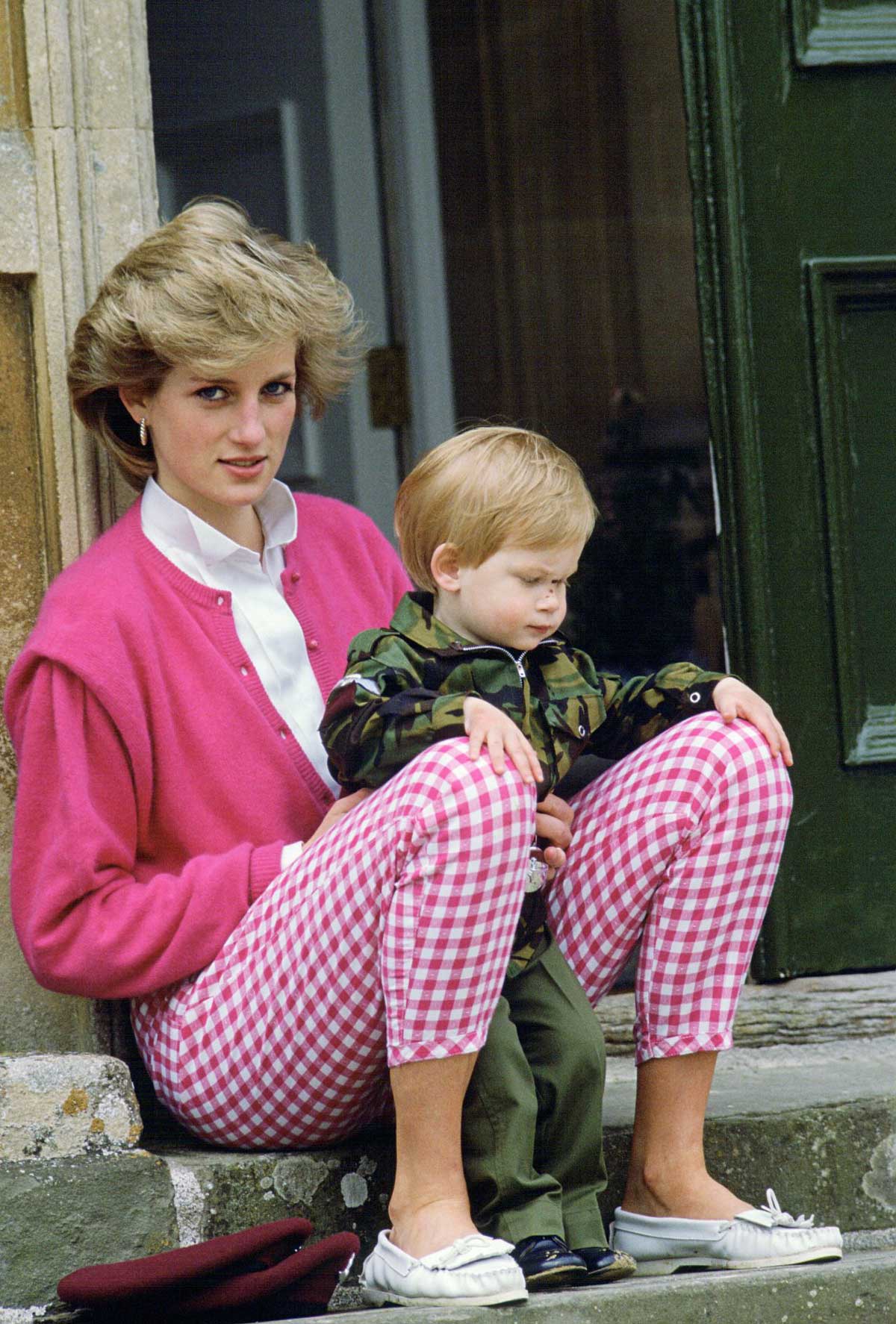Princess Diana in a bright pink cardigan and checkered pants sits on stone steps with a small child in a camouflage jacket on her lap, both looking down, near a green door.