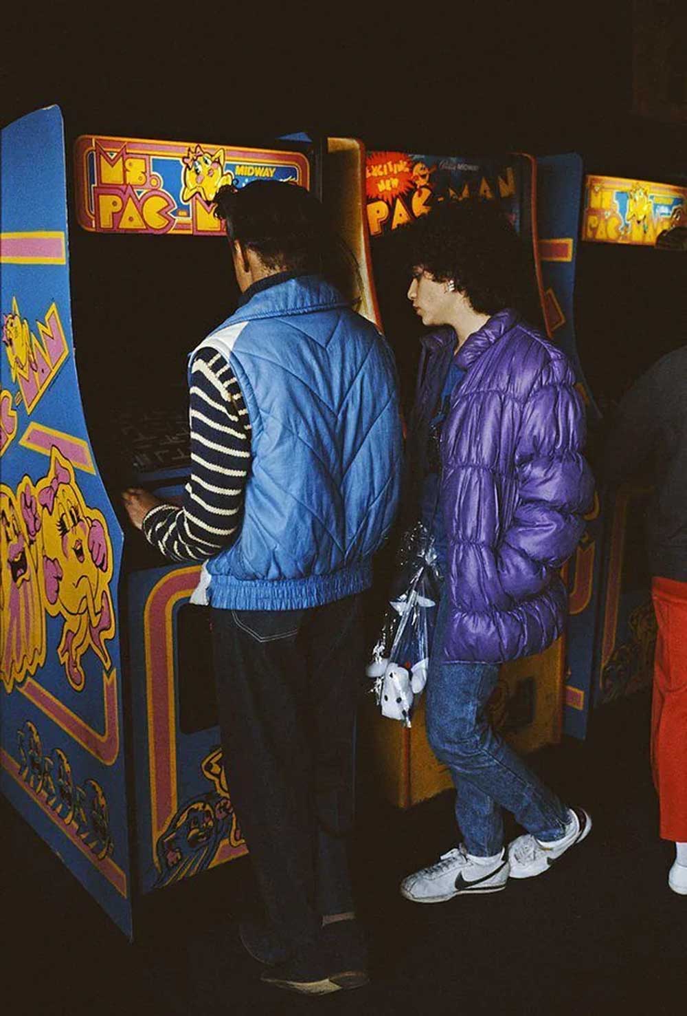 Two people wearing colorful jackets stand at a Ms. Pac-Man arcade machine, one playing the game while the other watches. The scene has a retro vibe with bright, classic arcade artwork and dim lighting.