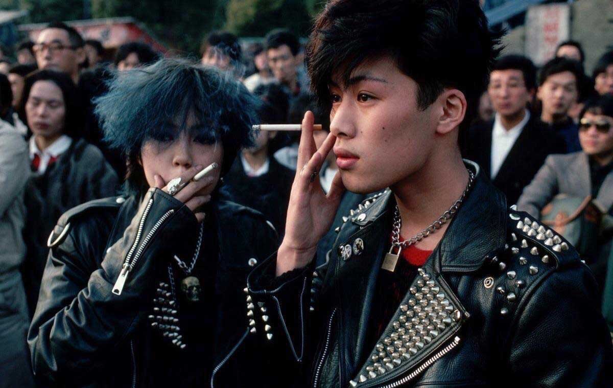 Two young people in spiked leather jackets stand in a crowd. One has blue hair and holds a cigarette, while the other touches their face. Both have edgy, punk-inspired fashion and serious expressions.