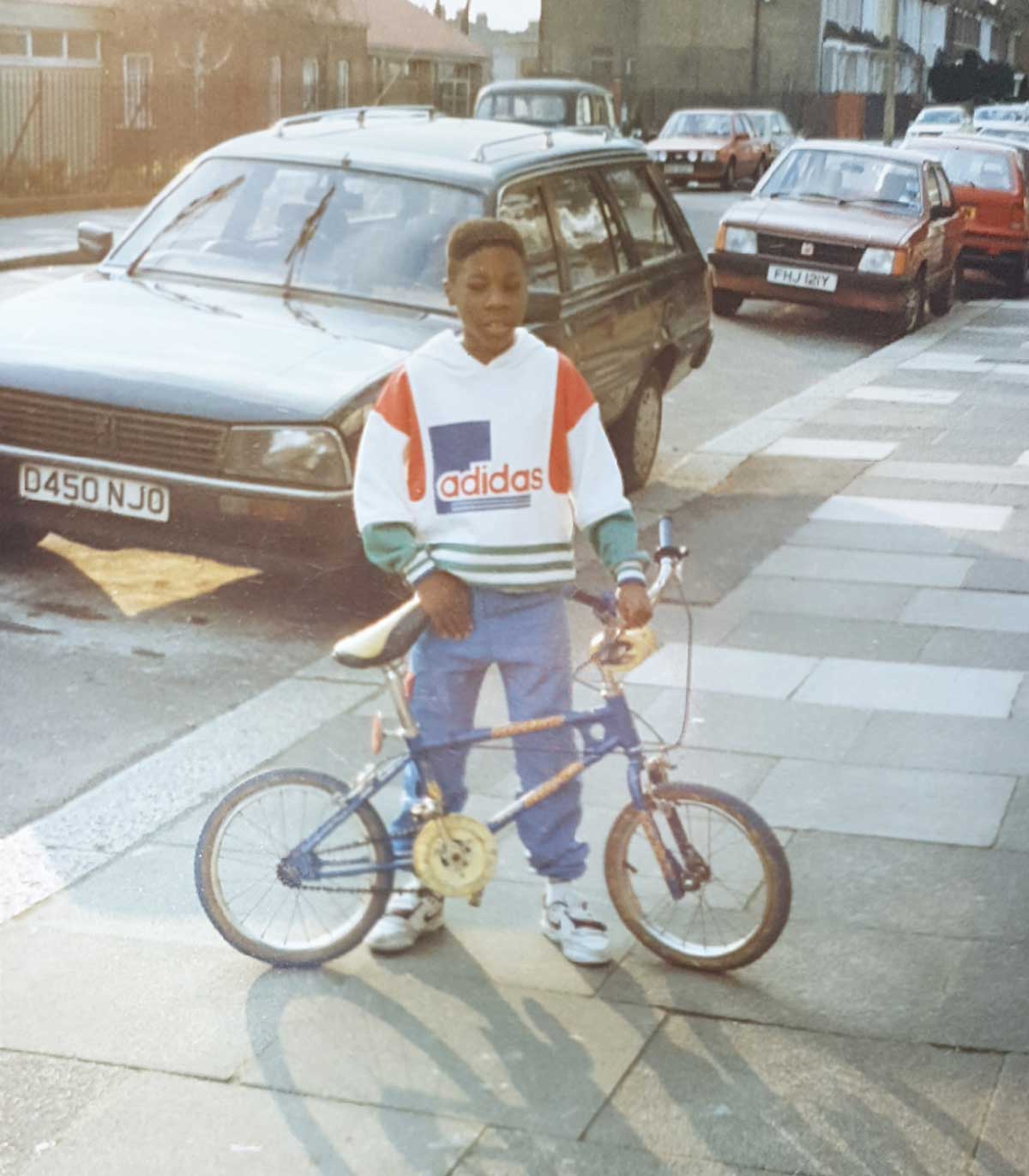 A boy stands on a sidewalk holding a BMX bike. He wears a white Adidas sweatshirt, blue pants, and sneakers. Parked cars and houses line the street in the background on a sunny day.
