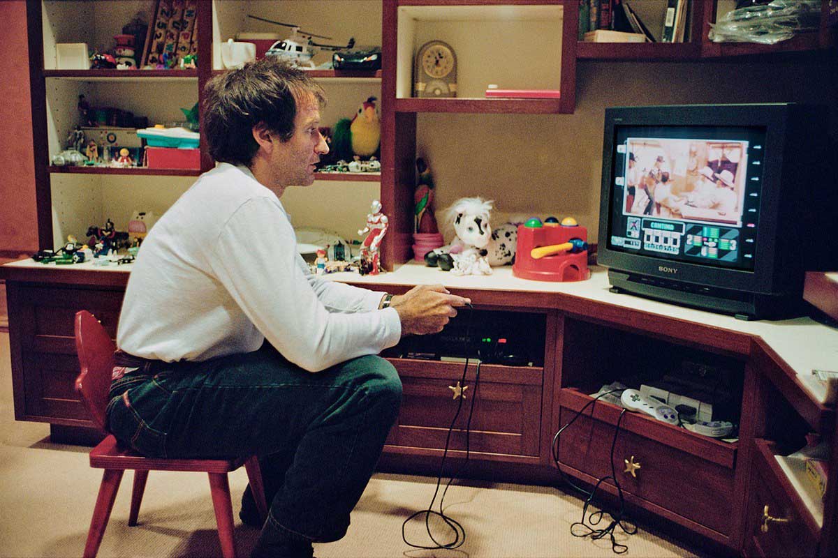 A man sits on a small red chair in a room, playing a video game on a vintage CRT TV. Shelves behind him hold toys, books, and collectibles. Game controllers and a console are visible on the entertainment unit.