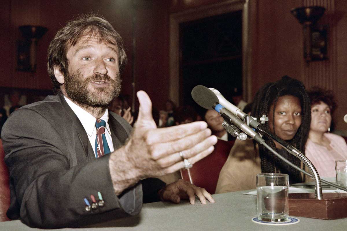 A bearded man in a suit gestures while speaking into a microphone at a hearing. A woman with long hair sits beside him, and other people are seated in the background. Glasses of water are on the table.