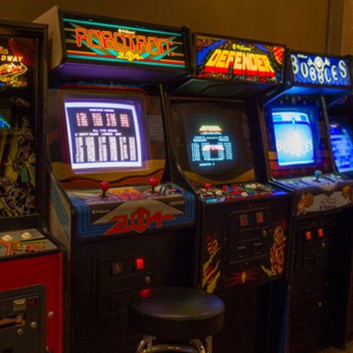 A row of vintage arcade game machines, including Robotron 2084, Defender, and Bubble Bobble, with illuminated screens and colorful artwork, stands beside a black stool in a dimly lit arcade.