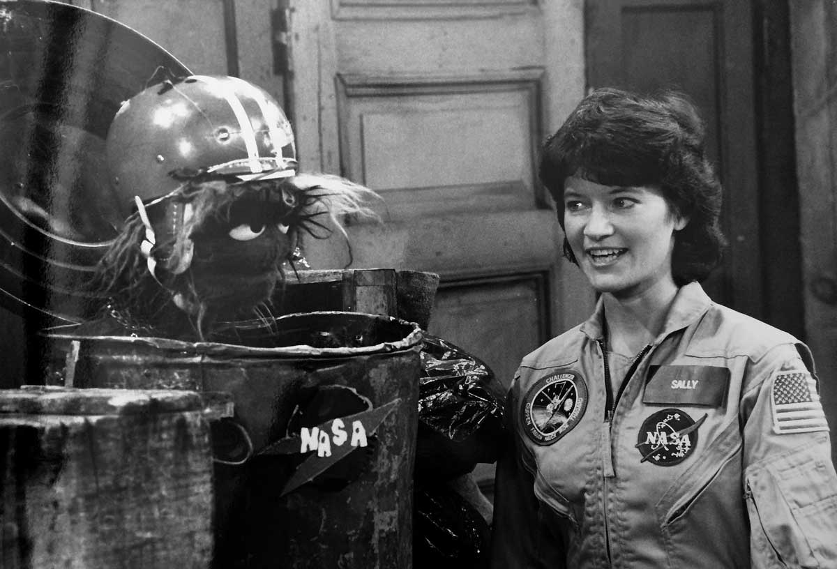 A woman in a NASA jumpsuit labeled Sally smiles at a puppet with a helmet emerging from a trash can labeled NASA. They appear to be on a set with wooden doors in the background.