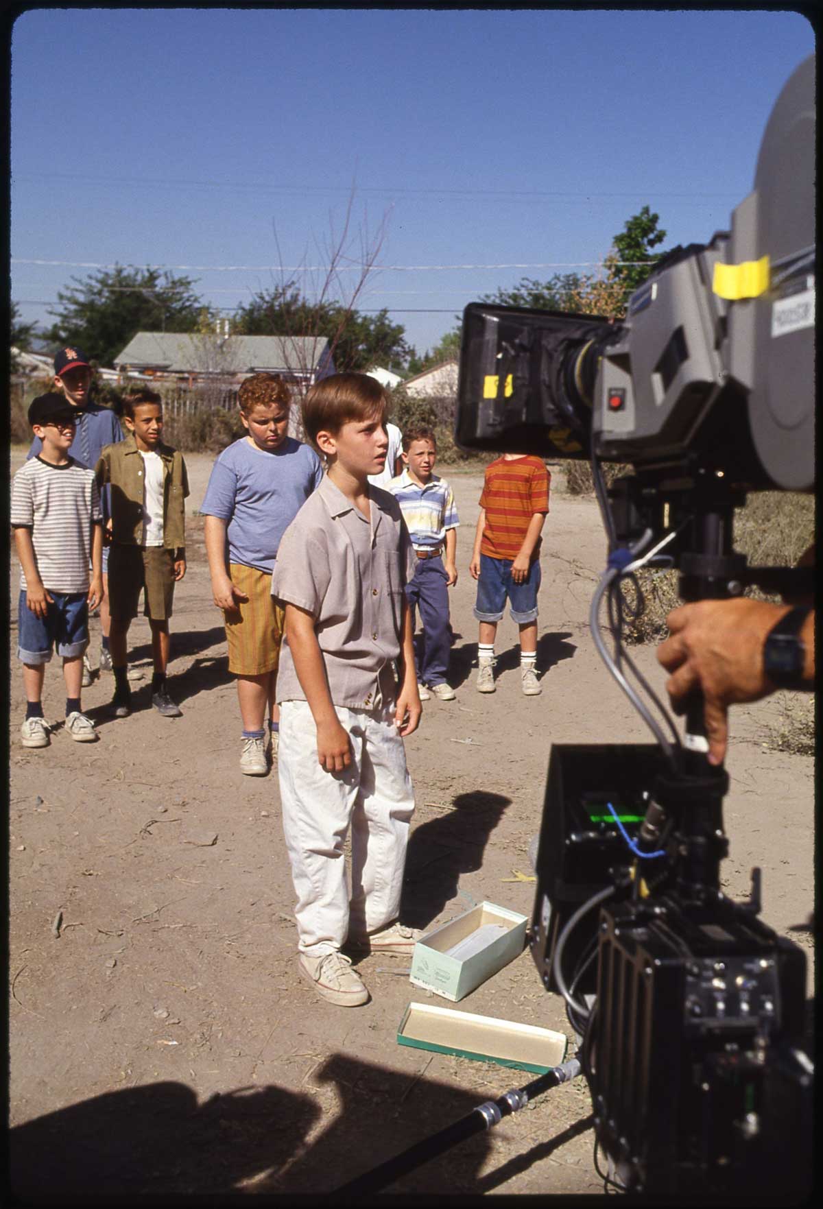 A group of young boys stands outdoors on a dirt path, with one boy in front facing a large film camera, suggesting a movie or TV scene being filmed in a neighborhood setting.