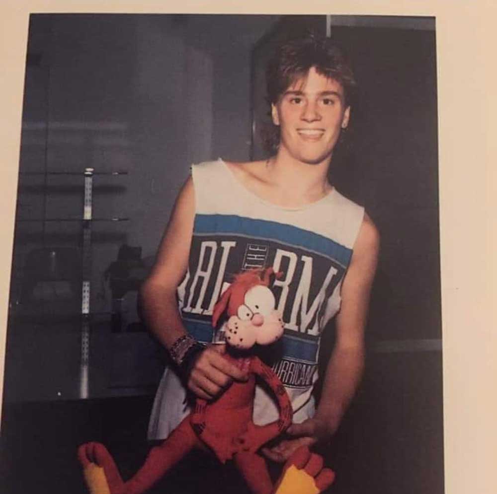 A young person with short, light brown hair wears a sleeveless The Alarm shirt and smiles while holding a plush toy of Garfield the cat in both hands.