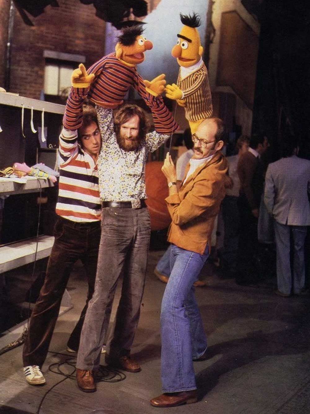 Three men backstage operate Bert and Ernie puppets from Sesame Street; two hold and control the puppets above their heads while the third gestures with a finger gun, all surrounded by stage equipment.