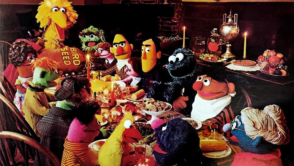The Sesame Street Muppets gather around a dinner table set with food, candles, and a silver teapot. Big Bird holds a bag of bird seed, and everyone appears to be enjoying a festive meal together.