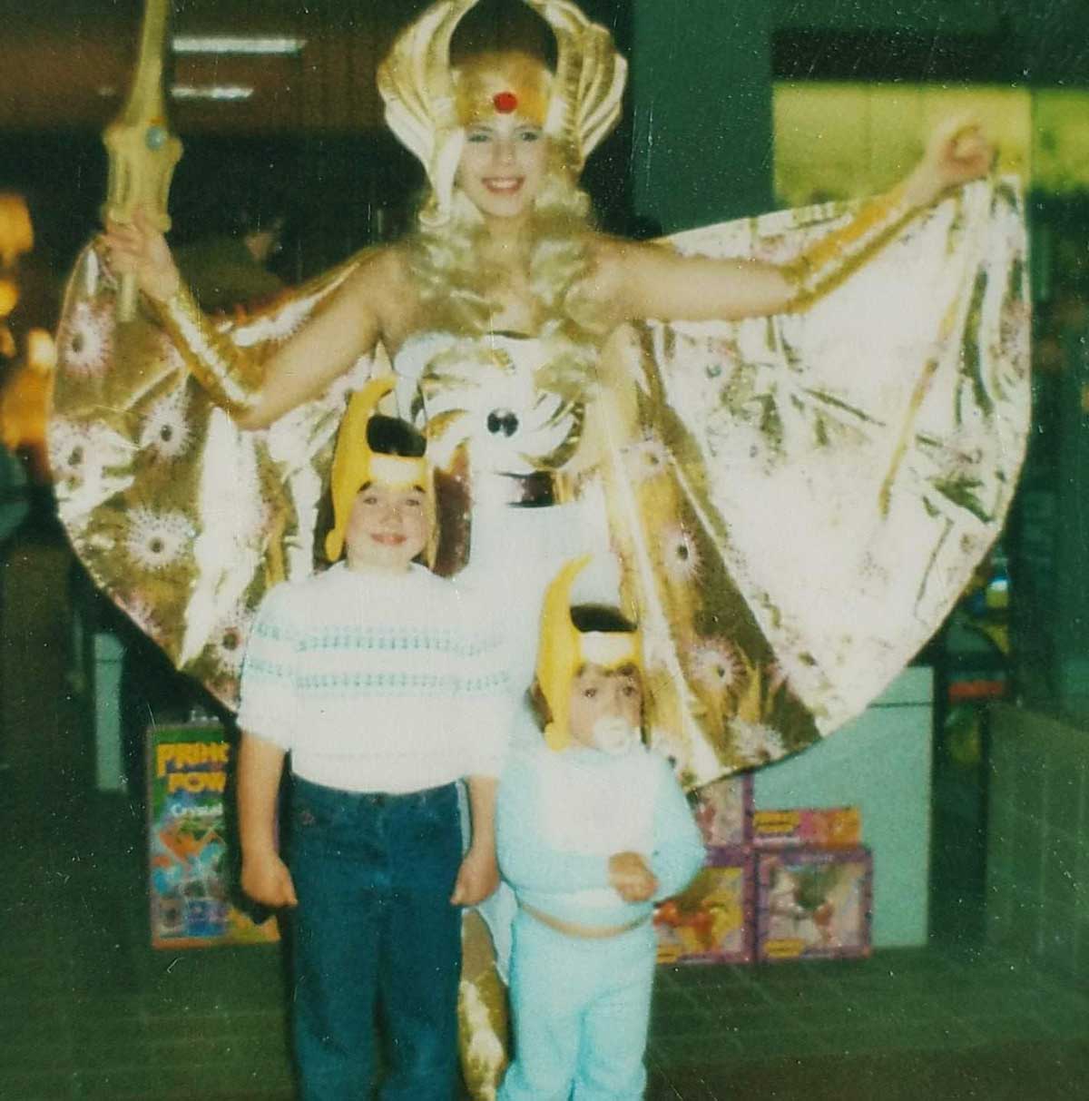 A woman dressed as She-Ra poses with two children wearing bird hats and sweaters in front of toy store shelves filled with colorful boxes.