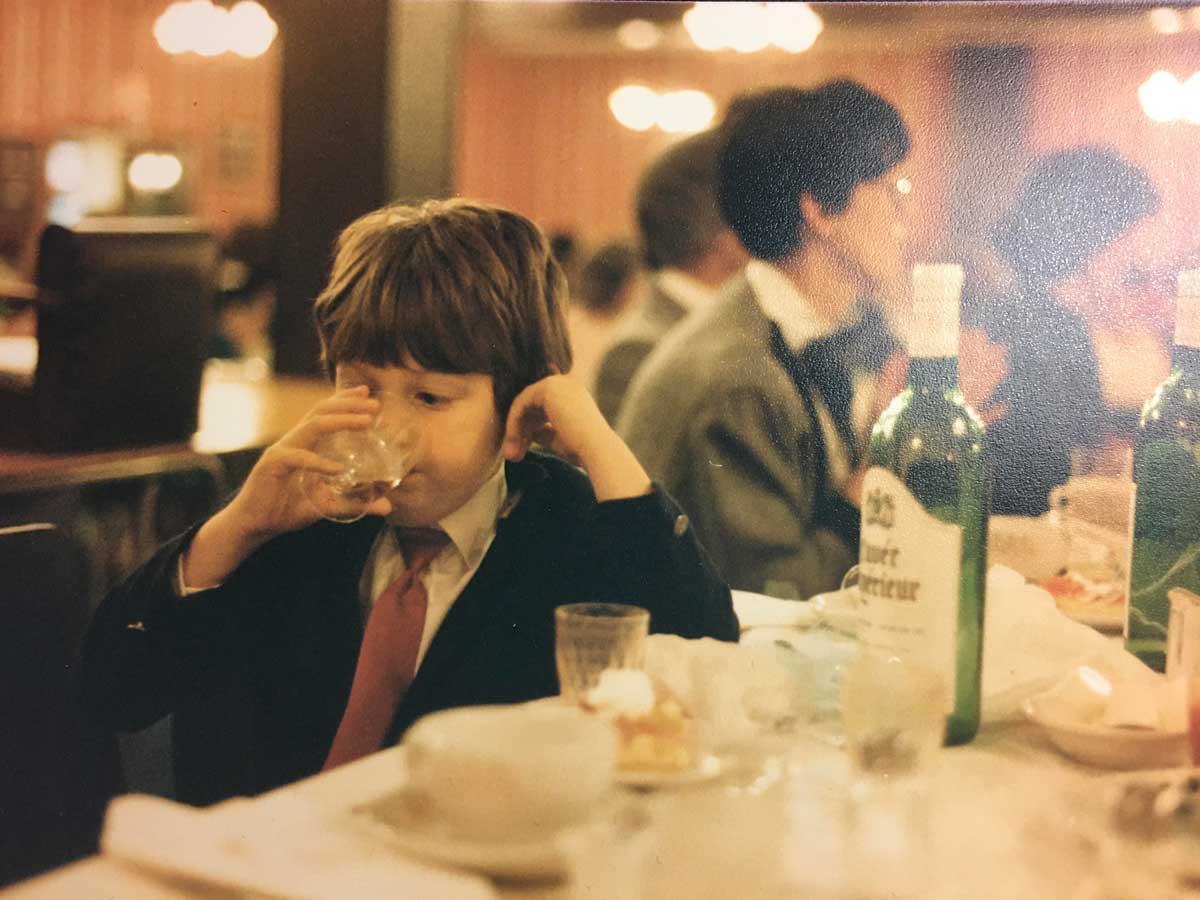 A young child in a suit and red tie sits at a dining table, sipping from a glass. The table is set with dishes, a piece of cake, and green wine bottles. Blurred adults sit in the warmly lit background.