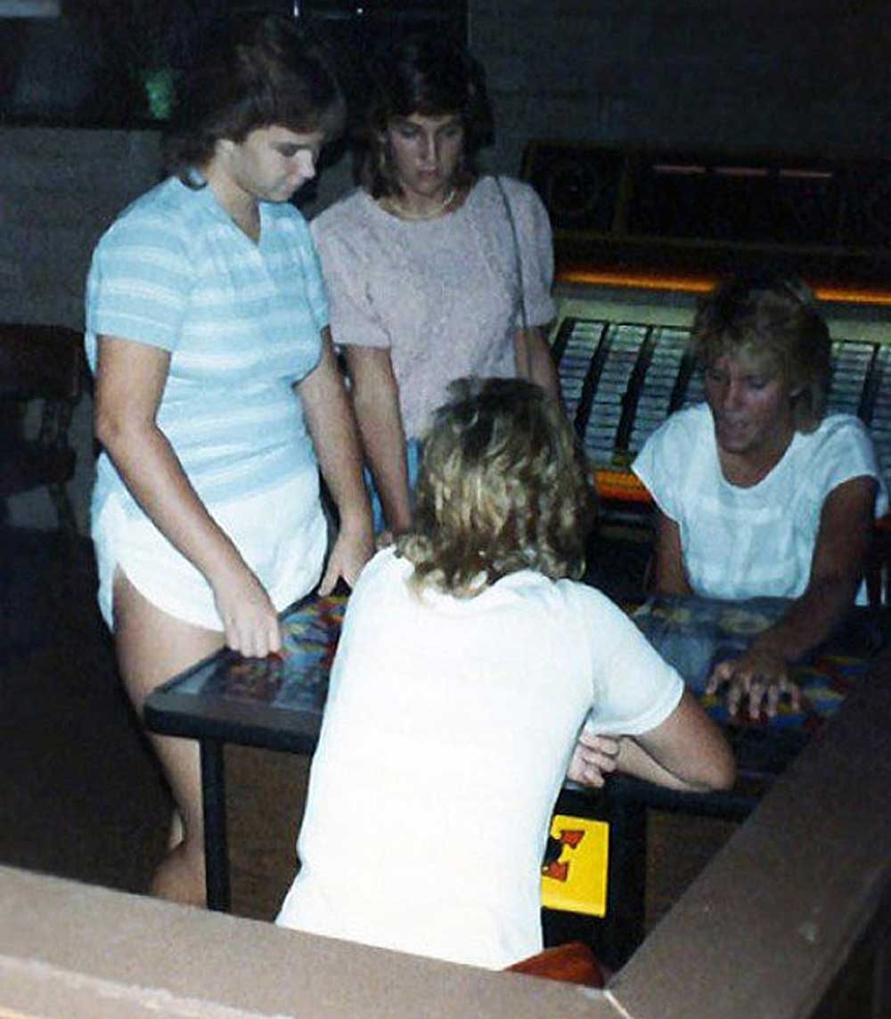Four young women stand and sit around a vintage tabletop arcade game, concentrating on the screen. A jukebox is visible in the background. The setting appears to be indoors, possibly in a game room or arcade.