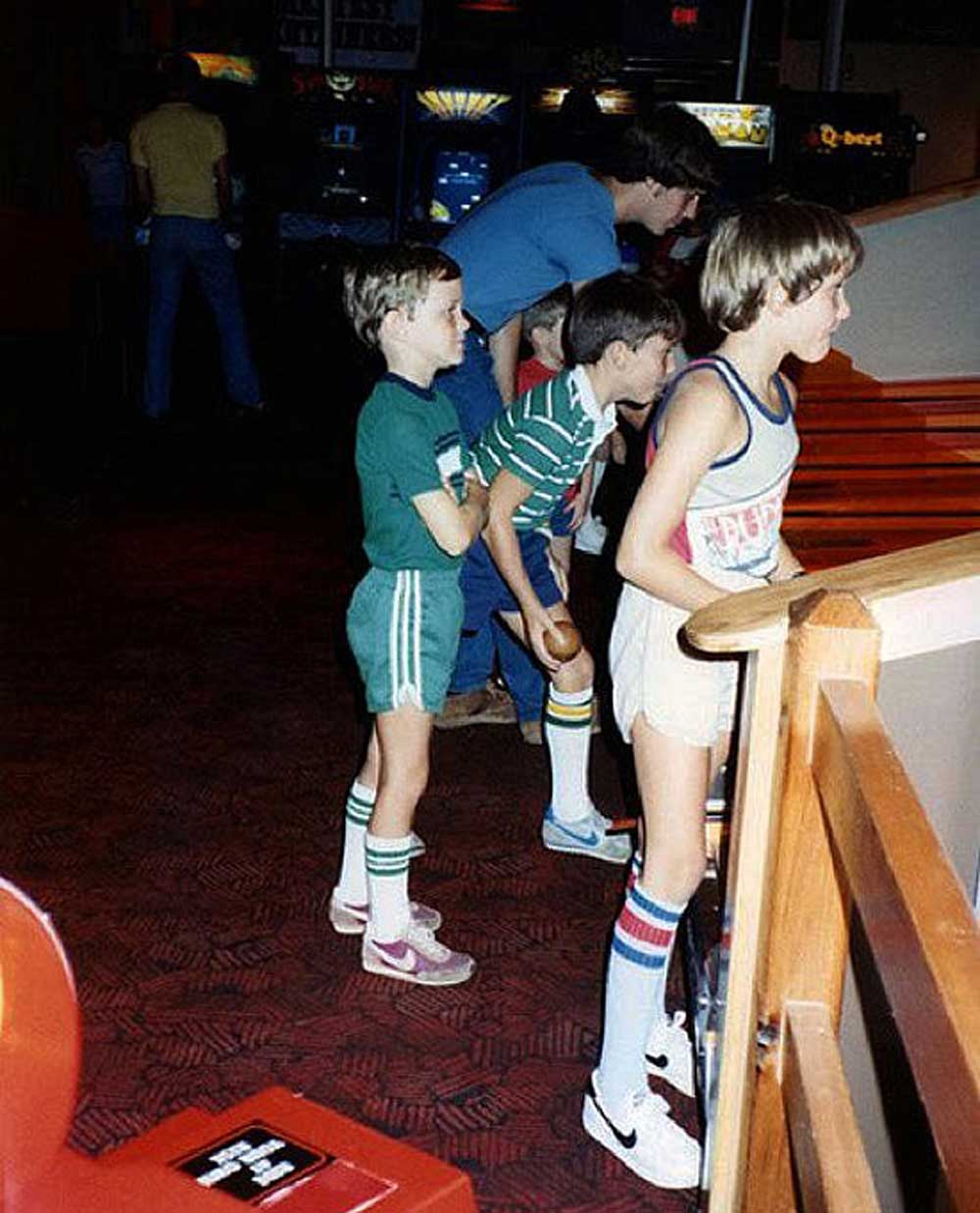 Three boys in retro athletic clothing and knee-high socks stand in line at an arcade, focused on games ahead. The background shows arcade machines and another person playing.
