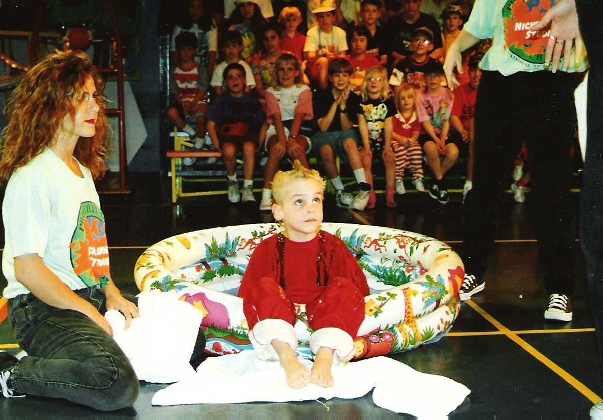 A young child in red sits in a colorful inflatable pool, surrounded by adults and towels, on a stage. An audience of children watches in the background.
