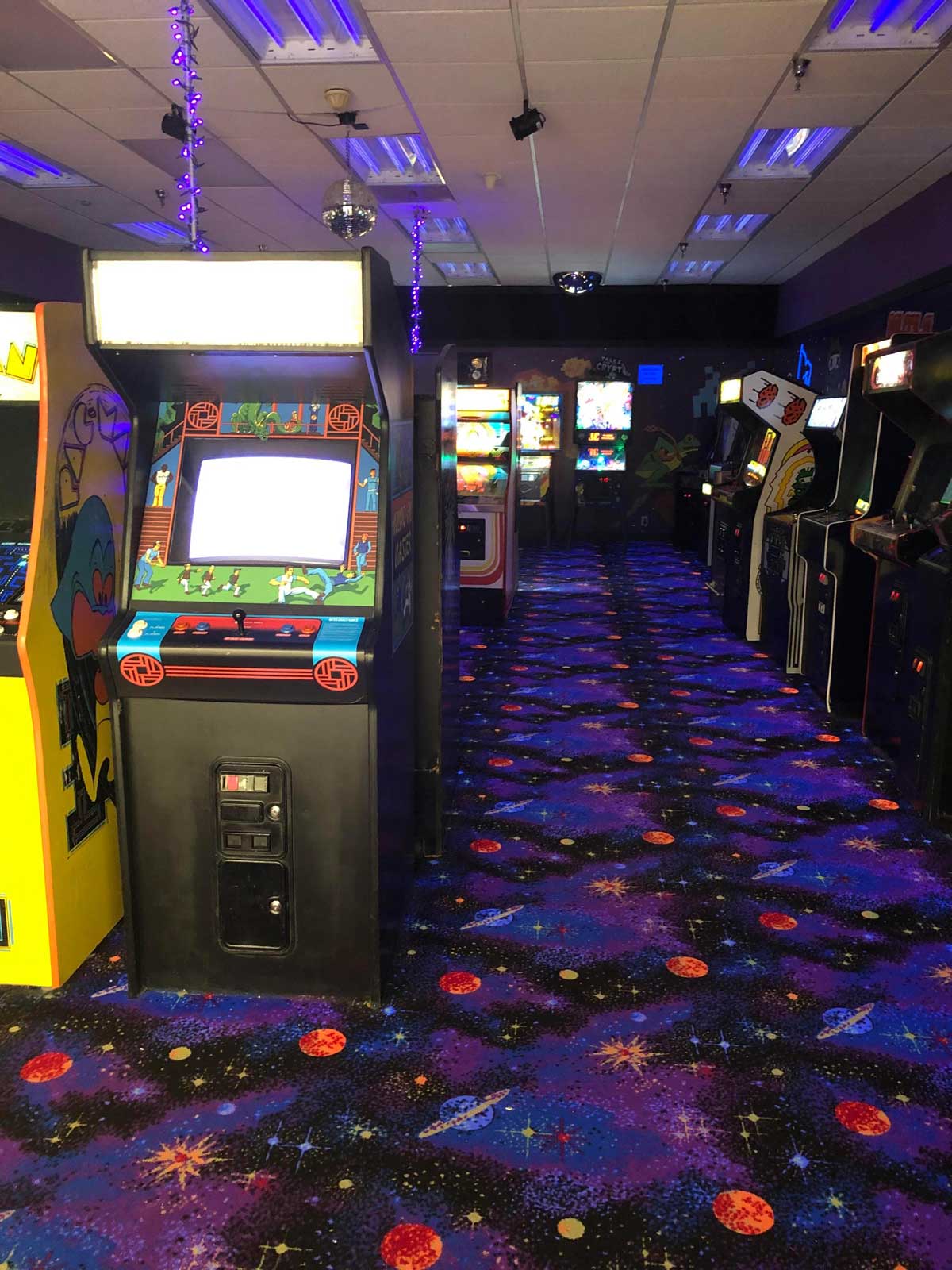 A retro arcade with rows of vintage arcade game machines and colorful, space-themed carpet covered in stars and planets. Purple string lights decorate the ceiling. The room is dimly lit.