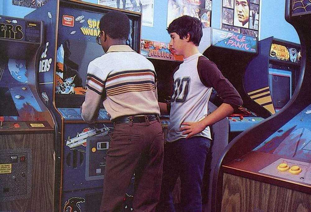 Two boys play arcade games in a retro-style arcade. One boy faces the machine, wearing glasses and a striped sweater, while the other stands beside him in a white and blue shirt. Posters and other arcade machines are in the background.