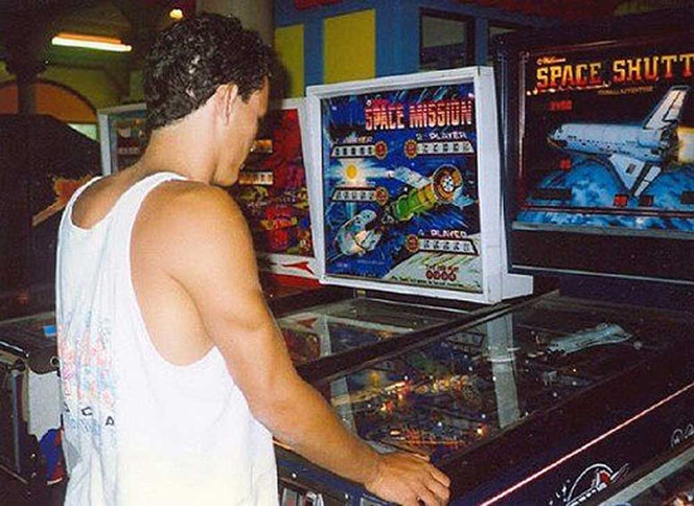 A man in a white tank top plays a Space Mission pinball machine in an arcade, with another pinball machine called Space Shuttle next to him. Bright colors and arcade decor are visible in the background.