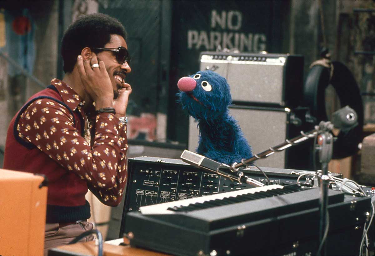 Stevie Wonder smiles while talking to Grover, a blue Sesame Street puppet, in a music studio with a keyboard and microphones in front of them. A No Parking sign is visible in the background.