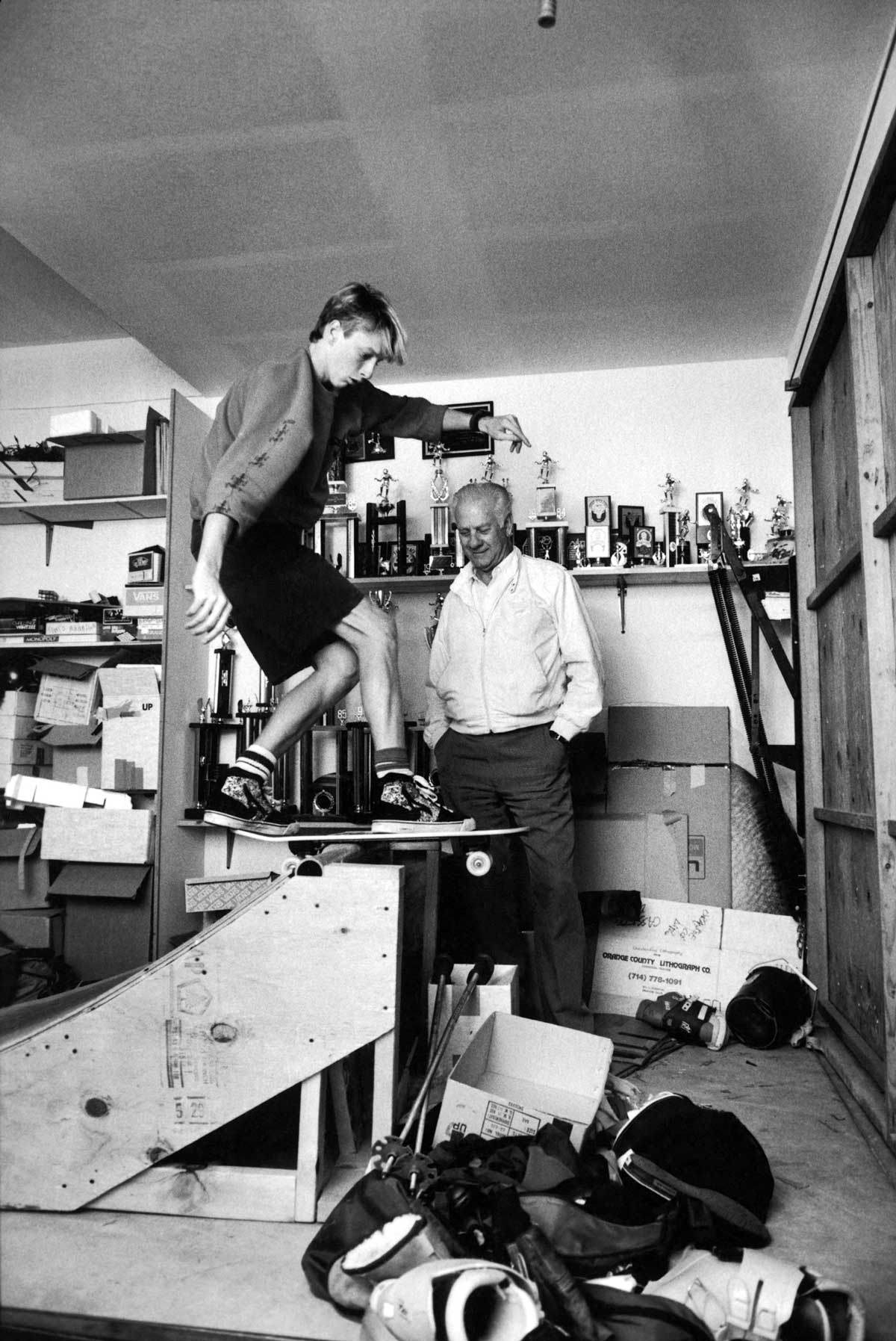Tony Hawk balances on a homemade wooden skateboard ramp indoors, while his dad Frank Hawk stands nearby smiling. The room is cluttered with boxes and trophies displayed on shelves in the background.