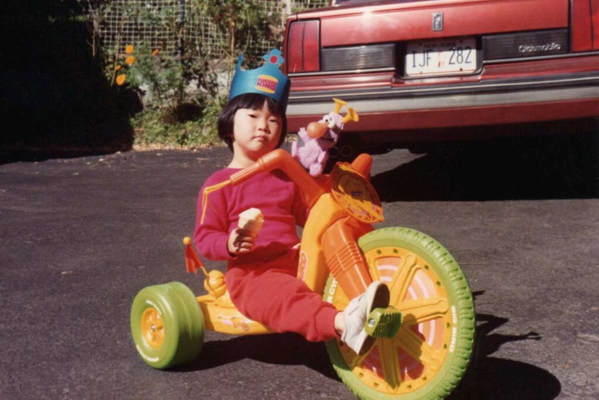 A young child wearing a paper crown and red clothes sits on a colorful tricycle, holding an ice cream cone. A toy is attached to the handlebars, and a red car is parked in the background.