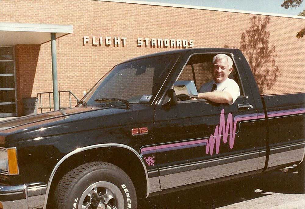 A man smiles while sitting in the drivers seat of a black pickup truck with purple graphics, parked in front of a building labeled FLIGHT STANDARDS.